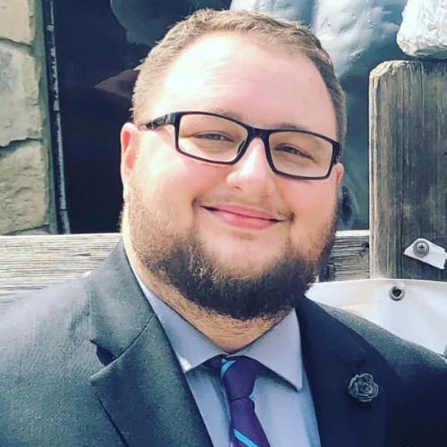 A smiling man with glasses and a beard wearing a dark suit and purple tie.